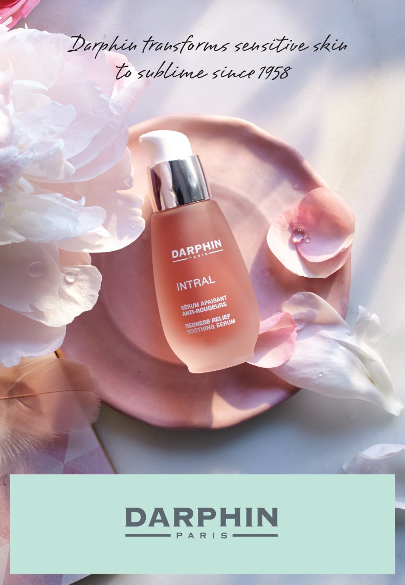 A bottle of Darphin Intral redness relief soothing serum placed on a pink plate surrounded by white and pink flower petals, with text indicating the product's benefits and a Darphin Paris logo below.