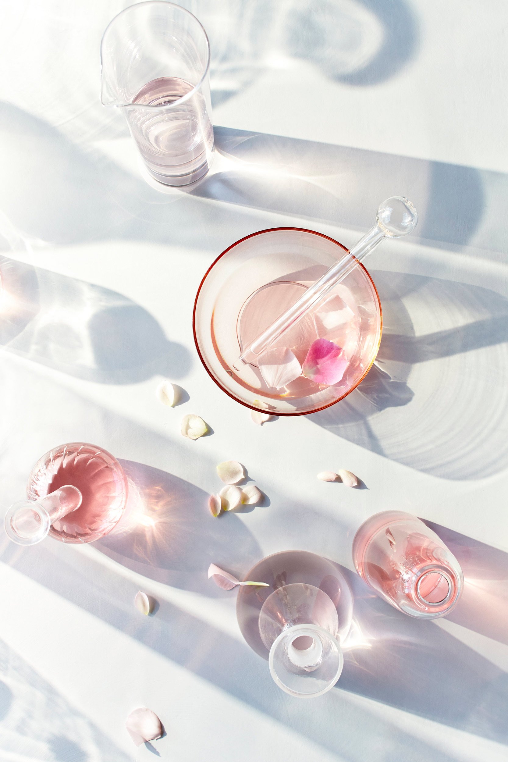 Several pink and clear glass vessels, some with flowers or petals, arranged on a white surface with shadows and scattered flower petals.